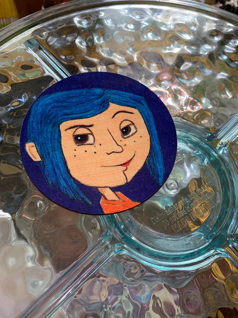 May include: A hand-painted wooden round coaster featuring a cartoon portrait of a girl with blue hair and a red shirt. The background is a dark blue color.
