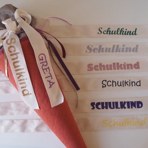 Könnte beinhalten: Ein roter und weißer Stoffkegel mit dem Namen "GRETA" bestickt. Der Kegel ist mit einem goldenen Band verziert, auf dem "Schulkind" steht. Es gibt mehrere weiße Stoffstreifen mit dem Wort "Schulkind" in verschiedenen Farben darauf gedruckt.
