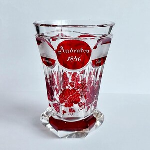 May include: A clear glass with a red base and rim, featuring a red circular design with the text "Andenten 1846". The glass is decorated with red grape vine patterns. The glass has a faceted base.