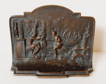 May include: A bronze bookend depicting a scene from Greek mythology. The bookend features a group of figures in a classical setting, including a man wrestling another man. The bookend is detailed and intricate, with a textured surface.