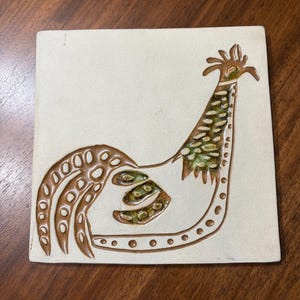 May include: Square ceramic tile with a rooster design. The rooster is depicted in brown and green against a cream-colored background. The design features a stylized rooster with a long neck and tail.