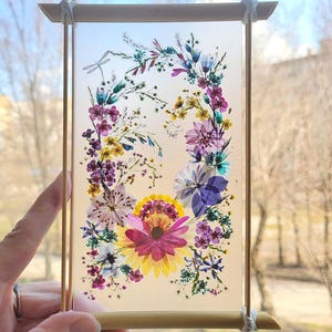 May include: A rectangular floral art piece featuring a vibrant arrangement of pressed flowers in shades of purple, yellow, and white. The flowers are arranged in a circular pattern within a clear panel, framed by a light brown wooden border. The piece is held by a hand.