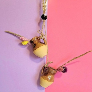 Op de afbeelding: Twee kleine bruine en beige keramische kannetjes met een bruin touwtje dat erboven hangt. De kannetjes zijn gevuld met gedroogde bloemen en hangen tegen een paarse en roze achtergrond.