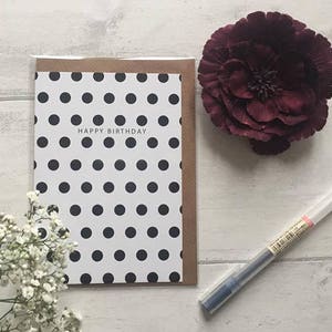 May include: A white card with black polka dots and the text "Happy Birthday" on a brown background. The card is next to a burgundy flower and a clear pen.