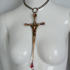 Puede incluir: Un collar con un colgante de daga dorado con detalles de piedras preciosas rojas. La daga está unida a una cadena con un anillo grande en la parte superior. La daga tiene una hoja larga y puntiaguda y un mango en forma de cruz.