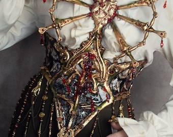 May include: A gold and red ornate corset with a heart design and dangling beads. The corset is worn over a white blouse with ruffled sleeves.