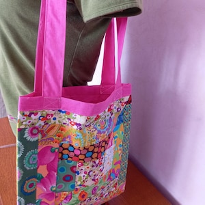 Peut inclure: Un sac fourre-tout présentant un motif patchwork composé de divers tissus aux imprimés floraux et géométriques colorés. Le sac est doté de anses en tissu rose vif et d'une bordure assortie sur le dessus, créant un contraste avec le corps multicolore.