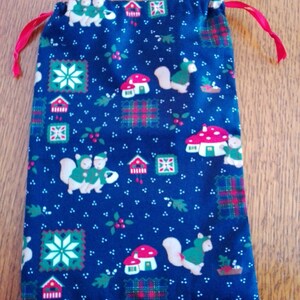 Peut inclure: Un sac bleu marine avec cordon de serrage orné d'un motif répété d'écureuils, de champignons, de maisons et de flocons de neige. Le sac a une fermeture à cordon de ruban rouge. Le tissu présente des accents rouges, verts et blancs.