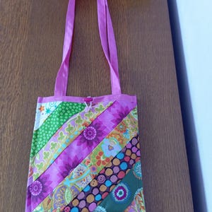 Peut inclure: Un sac fourre-tout rectangulaire en tissu avec un motif patchwork diagonal composé de divers imprimés colorés, notamment des fleurs, des pois et des formes géométriques dans des tons de rose, vert, jaune et bleu, avec une anse et un bouton rose.