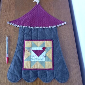 Peut inclure: Une tenture murale matelassée avec une base festonnée gris foncé et une section de toit bordeaux ornée d'une bordure florale. Un bloc central carré présente un motif d'étoile jaune et vert avec un appliqué de poulet rouge. Un stylo est présent.