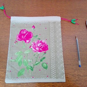 Peut inclure: Un sac en tissu beige avec un imprimé de roses roses vives. Le sac a une bordure blanche avec un motif à pois et un cordon de serrage rose avec des détails verts. Un stylo et des ciseaux sont à côté du sac.