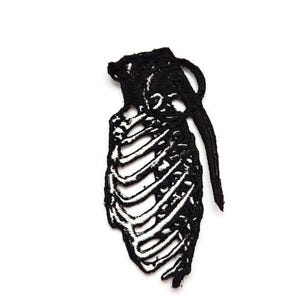 Pode incluir: Patch bordado preto em forma de granada com um design de esqueleto. O patch apresenta um contorno detalhado da forma da granada, com a caixa torácica do esqueleto visível. O patch está em um fundo branco.