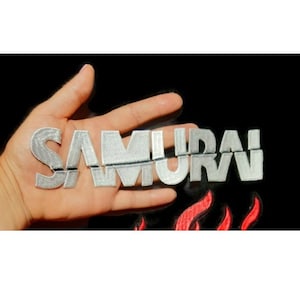May include: Embroidered patch with the word "SAMURAI" in silver thread. The patch is held in a hand against a black background with red flames at the bottom. The patch is likely for clothing or accessories.