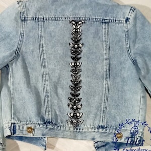Könnte beinhalten: Eine hellblaue Jeansjacke mit einer Reihe von schwarzen und weißen Schmetterlingsapplikationen auf dem Rücken. Die Applikationen haben die Form von Schmetterlingen mit Totenköpfen in der Mitte. Die Jacke hat einen klassischen Jeans-Stil mit Knopfleiste und zwei Taschen auf der Vorderseite.