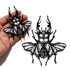 Könnte beinhalten: Zwei schwarze, gestickte Bügeleisen-Aufnäher von Hirschkäfern mit weißen Flügeln. Der größere Käfer ist etwa 7,5 cm breit und der kleinere etwa 5 cm breit.