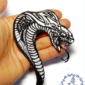 May include: Black and white embroidered iron-on patch of a snake's head with a forked tongue. The patch is held in a hand. The text "Taiss Embroidery" is visible in a wreath.