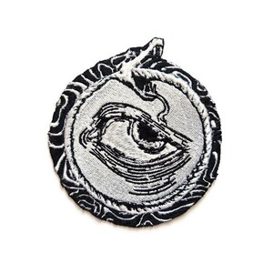 May include: Embroidered patch featuring a stylized eye design. The patch has a circular shape with a black and white color scheme. The eye is in the center, surrounded by a detailed border. The patch is likely for clothing or accessories.