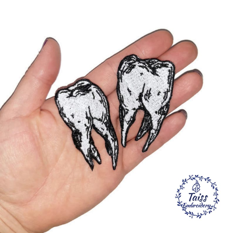 May include: Two white embroidered iron-on patches of teeth. The patches are on a hand, with the teeth facing outwards.