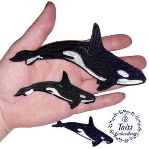May include: Two black and white embroidered orca whale patches. The larger patch is about 10 cm long and the smaller patch is about 5 cm long. The patches are on a hand.