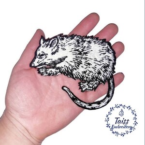 May include: Black and white embroidered iron-on patch of a sleeping possum. The patch is on a hand.