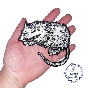 May include: Black and white embroidered iron-on patch of a possum. The possum is curled up with its tail wrapped around its body. The patch is approximately 10cm wide.
