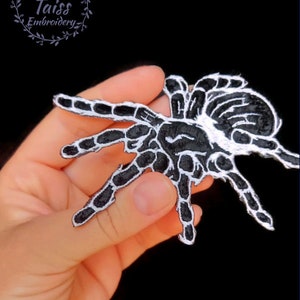 Peut inclure: Patch thermocollant brodé noir et blanc représentant une araignée avec un contour blanc. Le patch est tenu dans une main.