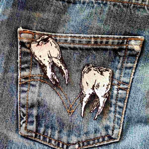 May include: A pair of white embroidered teeth sewn onto a blue denim pocket. The teeth are stitched in a realistic style.