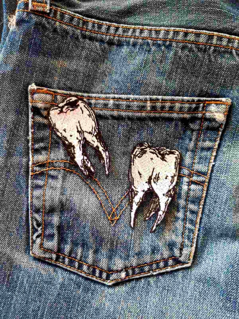 May include: A pair of white embroidered teeth sewn onto a blue denim pocket. The teeth are stitched in a realistic style.