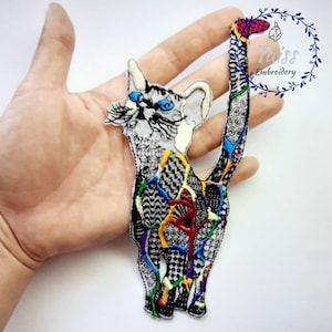 Puede incluir: Un parche bordado de colores para planchar con un gato sobre un fondo blanco. El gato está contorneado en negro y tiene una variedad de detalles coloridos en todo el diseño. La cola del gato está enroscada detrás de su espalda.