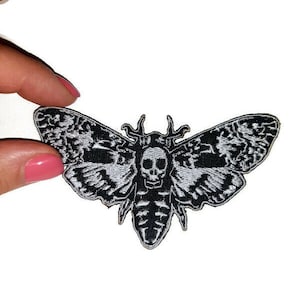 Puede incluir: Un parche bordado en blanco y negro con forma de polilla con un diseño de calavera en el cuerpo. Las alas de la polilla tienen patrones detallados. El parche se sostiene sobre un fondo blanco.