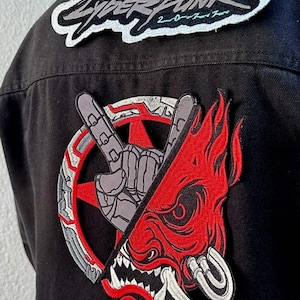 May include: A black denim jacket with three embroidered patches. The top patch is white and grey with the text "Cyberpunk 2077". The bottom two patches are a red and grey design with a hand making a rock and roll sign and a red demon face.
