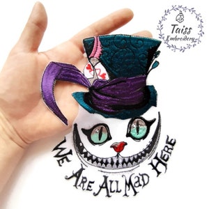Peut inclure: Écusson brodé représentant le Chat du Cheshire d'Alice au pays des merveilles. Le chat a un visage blanc, un large sourire et des yeux turquoise. Il porte un chapeau haut de forme violet et turquoise. Le texte "WE ARE ALL MAD HERE" est en dessous.