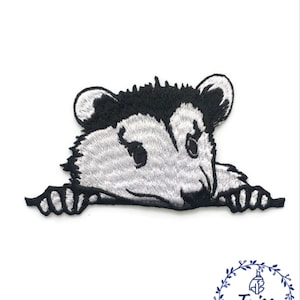 Puede incluir: Parche bordado para planchar de un opossum de dibujos animados gris y negro asomándose por encima de una superficie. El opossum tiene la nariz negra y los ojos negros.