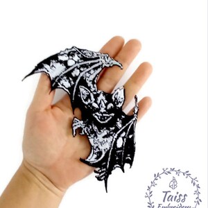 May include: Black and white embroidered iron-on patch of a bat with wings spread. The bat has a detailed design with a menacing expression. The patch is perfect for adding a spooky touch to clothing, bags, or other accessories.