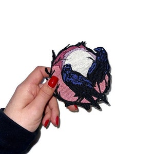 Puede incluir: Un parche bordado con dos cuervos negros posados en una luna rosa y blanca. El parche está rodeado por un contorno negro.