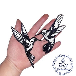 Puede incluir: Dos parches bordados de colibrí en blanco y negro. Los colibríes se miran, con las alas extendidas. Los parches están en una mano.