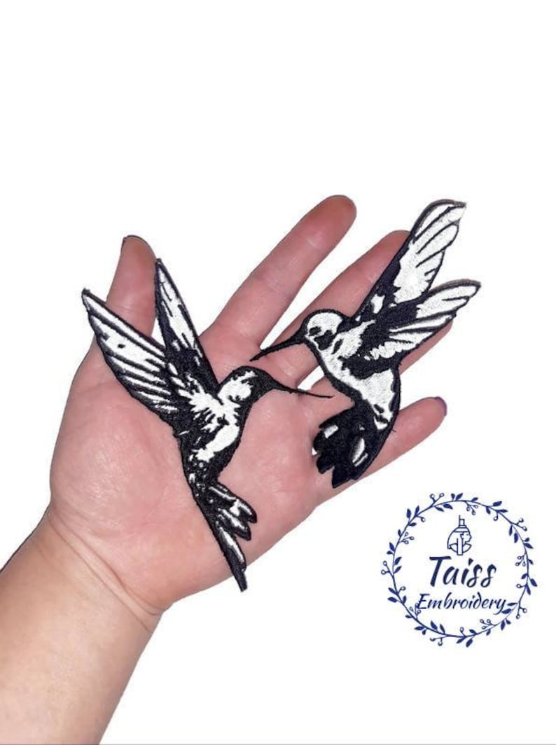 K&ouml;nnte beinhalten: Zwei gestickte Kolibri-Aufn&auml;her in Schwarz und Wei&szlig;. Die Kolibris schauen sich an, die Fl&uuml;gel ausgebreitet. Die Aufn&auml;her befinden sich auf einer Hand.