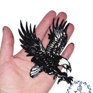 Puede incluir: Parche de hierro bordado en blanco y negro de un &aacute;guila en vuelo. El &aacute;guila se muestra con las alas extendidas y las garras extendidas. El parche mide aproximadamente 10 cm de ancho.