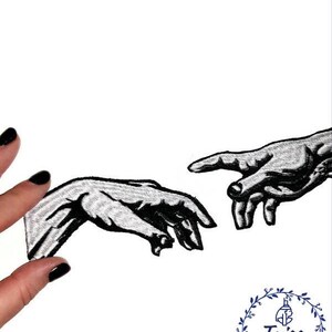 Pu&ograve; includere: Toppa ricamata in bianco e nero di due mani che si raggiungono, ispirata alla Creazione di Adamo di Michelangelo. La toppa &egrave; su uno sfondo bianco ed &egrave; tenuta da una mano con smalto nero.