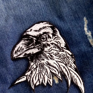 May include: A white embroidered raven patch with black stitching, ideal for adding a touch of gothic style to denim jackets, bags, or other fabric items.