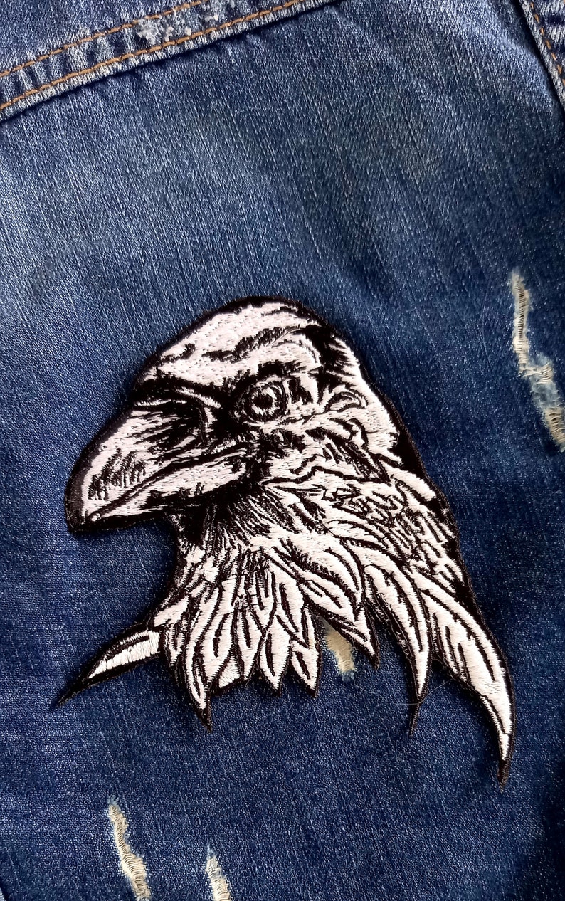 May include: A white embroidered raven patch with black stitching, ideal for adding a touch of gothic style to denim jackets, bags, or other fabric items.