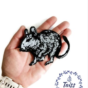 Puede incluir: Parche bordado en blanco y negro para planchar de un degu, un pequeño roedor, con un patrón de pelaje gris y blanco. El parche se sostiene en una mano.