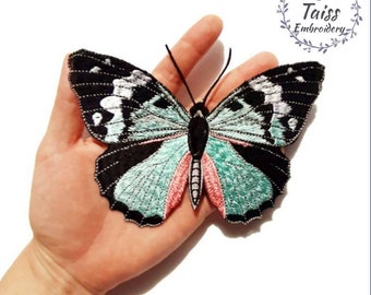 Könnte beinhalten: Ein gestickter Schmetterlings-Patch in Teal und Schwarz mit rosa Akzenten. Der Schmetterling wird auf einer Hand gezeigt, mit dem Text "Taiss Embroidery" in einem Kranz über dem Schmetterling.