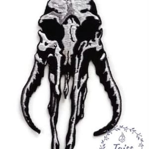 May include: Black and white embroidered patch of a skull with tusks. The skull is outlined in silver thread.