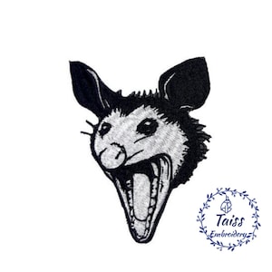 Peut inclure: Patch brodé noir et blanc d'un opossum la gueule ouverte, montrant ses dents. L'opossum regarde vers la droite. Le patch est sur un fond blanc.