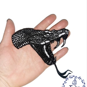 May include: Black and white embroidered iron-on patch of a snake's head with an open mouth and fangs. The patch is on a hand.