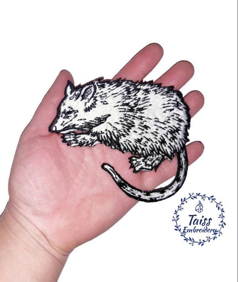 May include: Black and white embroidered iron-on patch of a sleeping possum. The patch is on a hand.