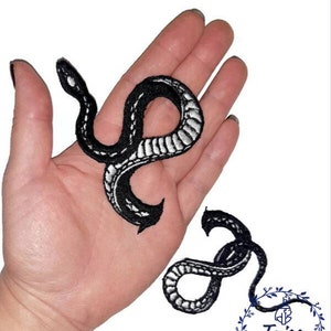 May include: Two black and white embroidered iron-on patches of snakes. The snakes are in a curled position. The patches are on a hand.