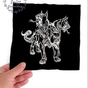 Puede incluir: Parche bordado blanco sobre tela negra que representa a un perro de tres cabezas, Cerbero, de la mitología griega.