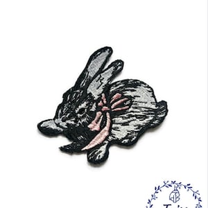 May include: Embroidered iron-on patch of a gray rabbit with a pink bow. The patch is perfect for adding a touch of whimsy to clothing, bags, or other fabric items.